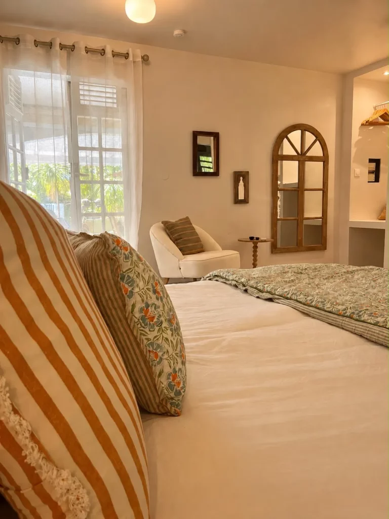 Chambre confortable du gîte Bois Banane avec lit double, miroir en bois sculpté et porte-fenêtre donnant sur la terrasse tropicale, Les Bois d'Orée à Sainte-Anne