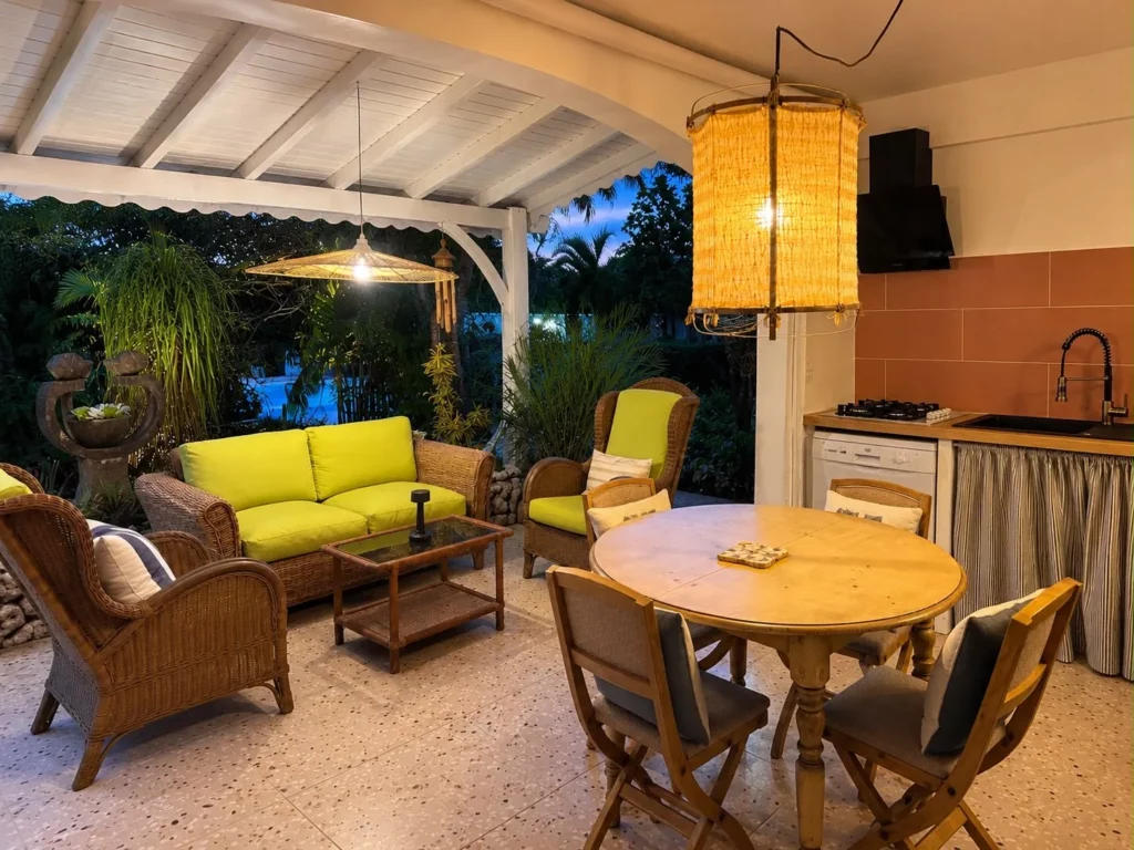 Terrasse couverte du gîte Bois Banane avec salon en rotin, table à manger et coin cuisine extérieure en soirée, Les Bois d'Orée, Sainte-Anne