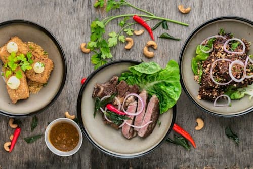 Trois assiettes de poisson et de viande garnies de légumes et d’herbes, sur une table en bois parsemée de piments et de noix de cajou.
