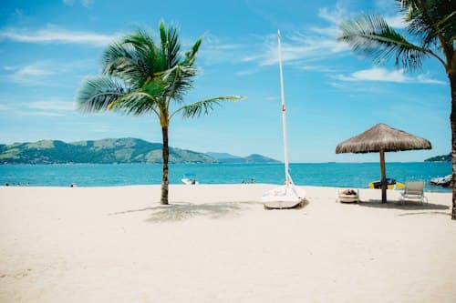 Plage de sable blanc bordée de palmiers, avec un petit voilier et des transats sous un parasol, face à une mer turquoise.