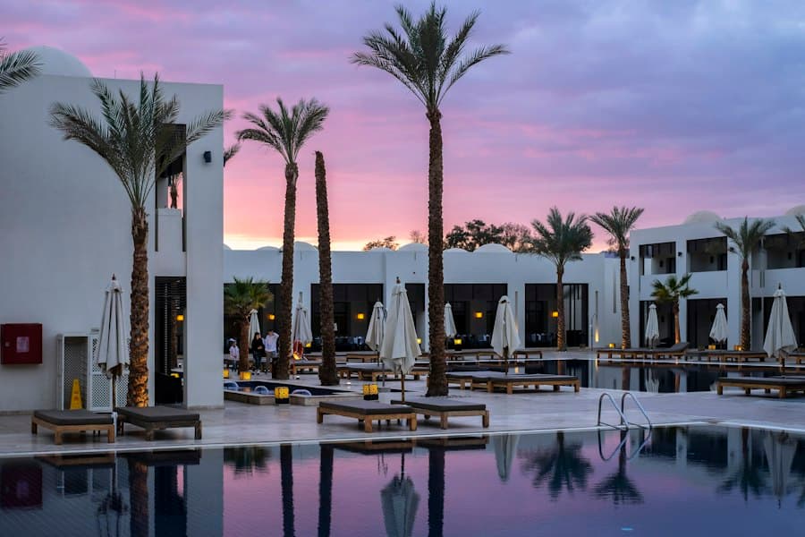 Piscine bordée de palmiers et de transats, entourée de bâtiments blancs sous un ciel rose au coucher du soleil.